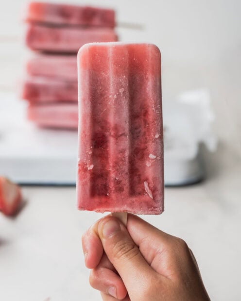 A hand holding a strawberry popsicle with more popsicles blurred in the background, 2K Mobile Wallpaper