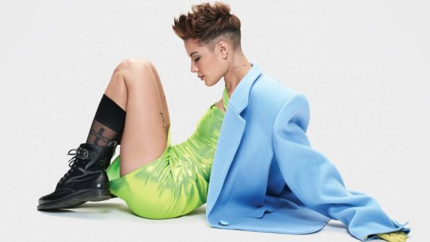 A side view of Halsey wearing a bright green dress and blue coat sitting against a white background