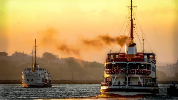 A ferry emits smoke while sailing during sunset over the sea with cityscape in the background, HD Desktop Wallpaper