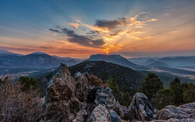 The sun sets behind the Rocky Mountain National Park with rocky foreground and forested hills, 2K Desktop Wallpaper