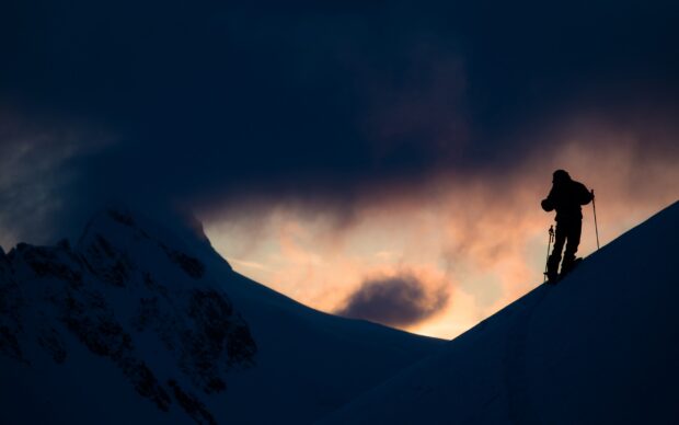 A skier is climbing a snowy mountain slope during sunset with dark clouds, 2K Desktop Wallpaper