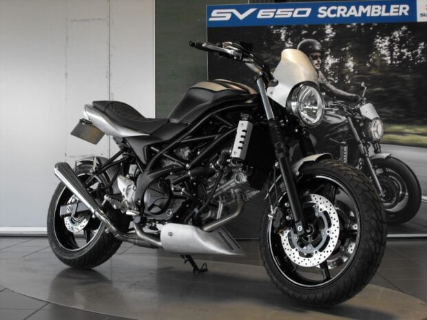 A black Suzuki SV650 scrambler motorcycle parked indoors with a promotional poster behind, HD Desktop Wallpaper