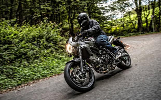 A rider on a Suzuki SV650 is leaning into a turn on a forested road with greenery in the background, 2K Desktop Wallpaper