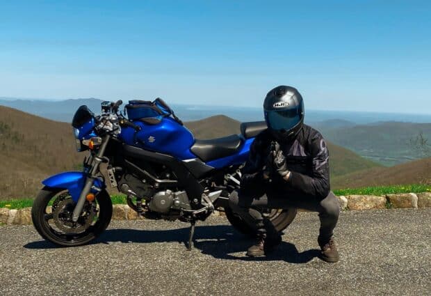 A rider wearing black gear poses next to a blue Suzuki Sv650 motorcycle on a mountain road, HD Desktop Wallpaper