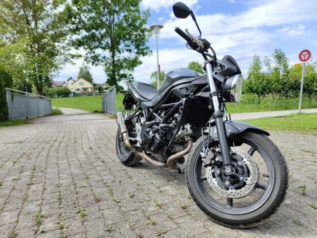 A Suzuki Sv650 motorcycle is parked on a paved path near green trees and houses, HD Desktop Wallpaper