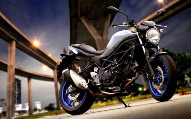 A Suzuki Sv650 motorcycle is parked near a modern bridge under a dark sky, 2K Desktop Wallpaper