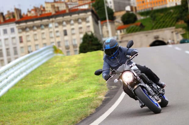 A rider in full gear is riding a Suzuki Sv650 on a sharp road curve near city buildings, HD Desktop Wallpaper
