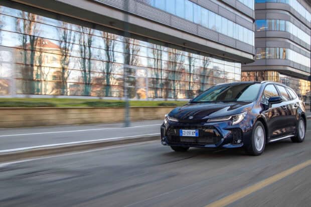 A Suzuki Swace is driving on an urban street with modern glass buildings, 2K Desktop Wallpaper