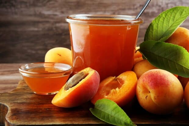 A jar of apricot jam is surrounded by fresh apricots and green leaves on a wooden surface, 2K Desktop Wallpaper