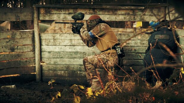 Two players engage in a tactical paintball game behind wooden cover, HD Desktop Wallpaper