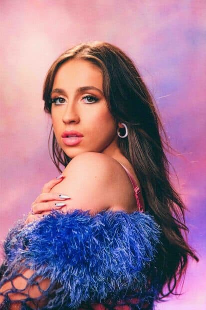 Tate Mcrae wallpapers showing her wearing a blue feathered outfit with a soft pink and purple background, HD Mobile Wallpaper