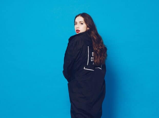 A woman with long brown hair and red lips wearing a black coat stands in front of a blue wall, HD Desktop Wallpaper