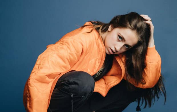 Tate Mcrae in a striking pose wearing an orange jacket against a blue background, HD Desktop Wallpaper