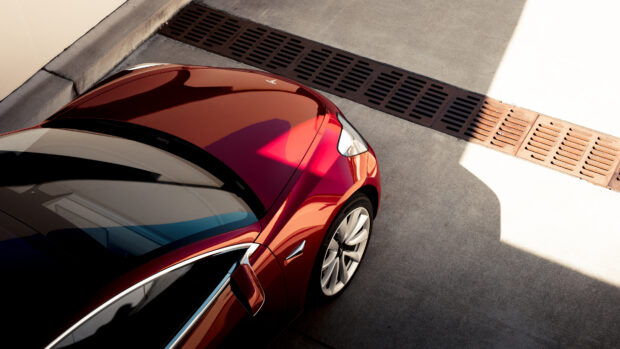 A red Tesla Model 3 is parked on a sunlit driveway with sharp shadows, 4K Desktop Wallpaper