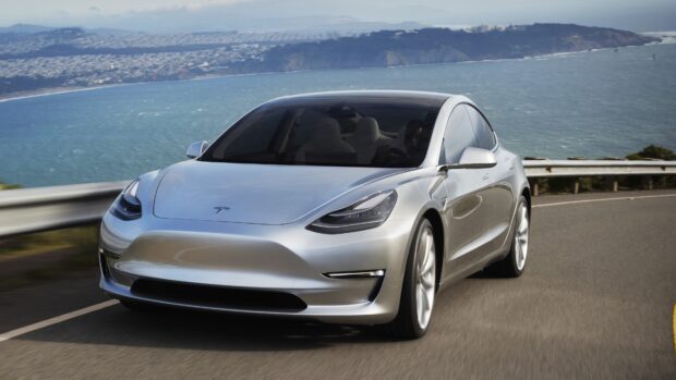 A silver Tesla Model 3 is driving along a coastal road with a view of the ocean and hills, 4K Desktop Wallpaper