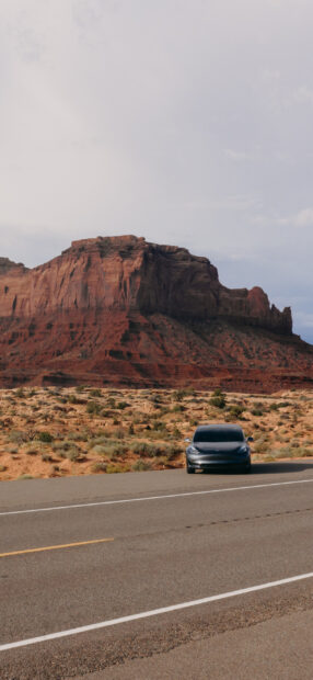 A Tesla Model 3 is parked on a desert road with rocky cliffs in the background, 2K Phone Wallpaper