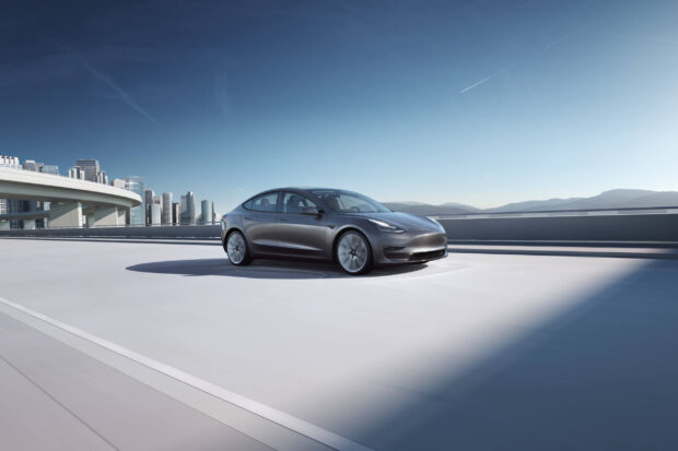 Tesla Model 3 is parked on a sunny urban highway with city buildings in the background, 2K Desktop Wallpaper