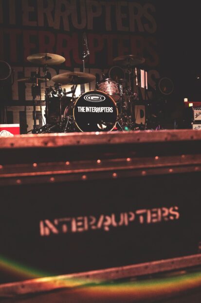 The Interrupters band drum set is displayed on a dimly lit stage with the band name prominently visible, HD Mobile Wallpaper