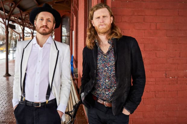 Two members of The Lumineers stand against a red brick wall, HD Desktop Wallpaper
