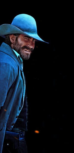 A man wearing a blue coat and hat smiles with a dark background in The Sisters Brothers, 2K Mobile Wallpaper