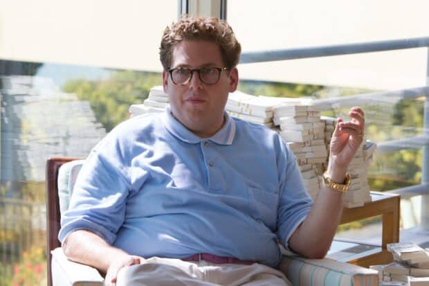 A man in a blue shirt sits in a chair surrounded by stacks of cash in this The Wolf Of Wall Street scene, 2K Desktop Wallpaper