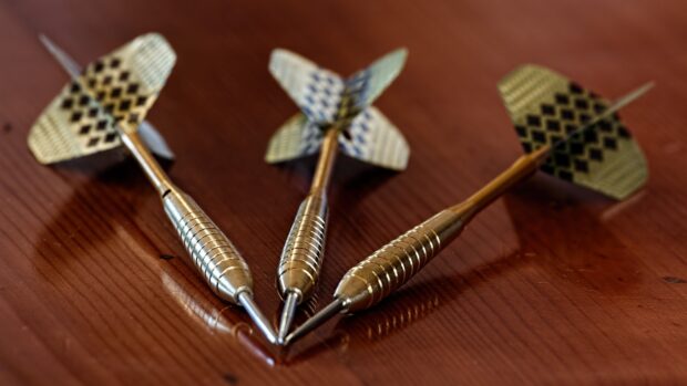 Three metal darts are arranged closely on a wooden tabletop featuring patterned flights, 4K Desktop Wallpaper