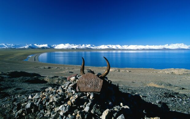 A rocky cairn with horns overlooks a serene lake and snowy mountains in the Tibetan Highlands, HD Desktop Wallpaper