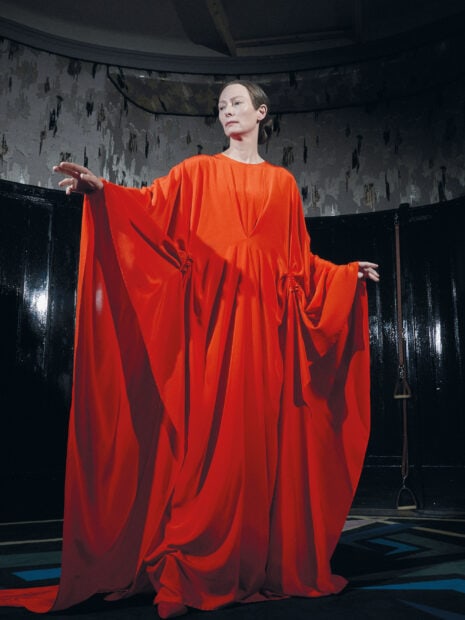 Tilda Swinton stands poised wearing a bright red flowing gown in a dimly lit room, 2K Portrait Desktop Wallpaper