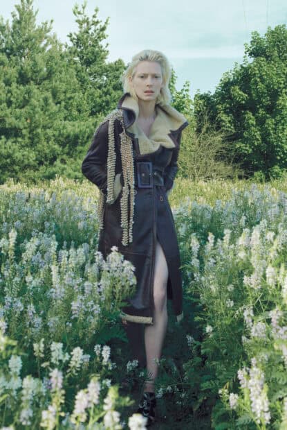 Tilda Swinton wearing a long coat while standing amidst blooming flowers and trees, HD Phone Wallpaper