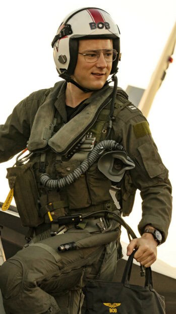 A Top Gun Maverick pilot is dressed in a flight suit and helmet, holding a bag next to a jet, HD Mobile Wallpaper