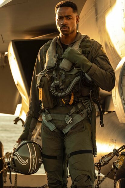 A pilot from Top Gun Maverick holds his helmet standing near a jet aircraft on the deck, 2K Mobile Wallpaper