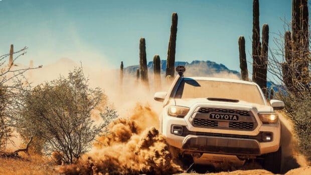 A Toyota rally raid vehicle kicks up dust while driving through a desert with cacti and mountains, 2K Desktop Wallpaper