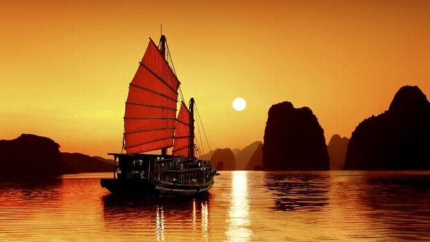 A traditional boat sails peacefully at sunset among limestone islands in Vietnam with warm orange skies reflecting on the water, 4K Desktop Wallpaper