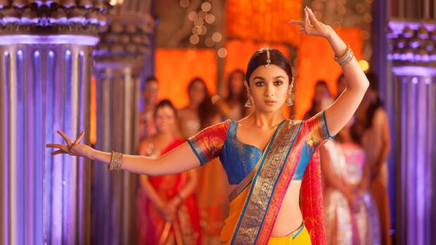 A woman in vibrant traditional attire performing a dance pose in a festive Bollywood scene, 4K Desktop Wallpaper