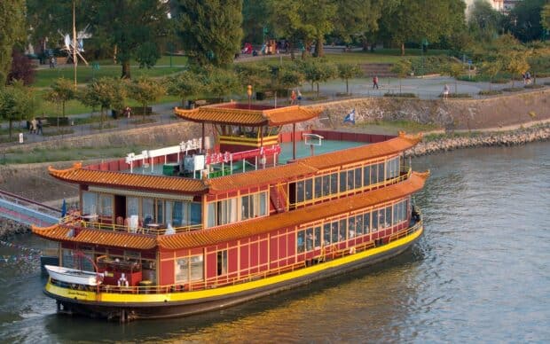 A traditional ferry with ornate design sails close to the riverbank with people walking nearby, HD Desktop Wallpaper