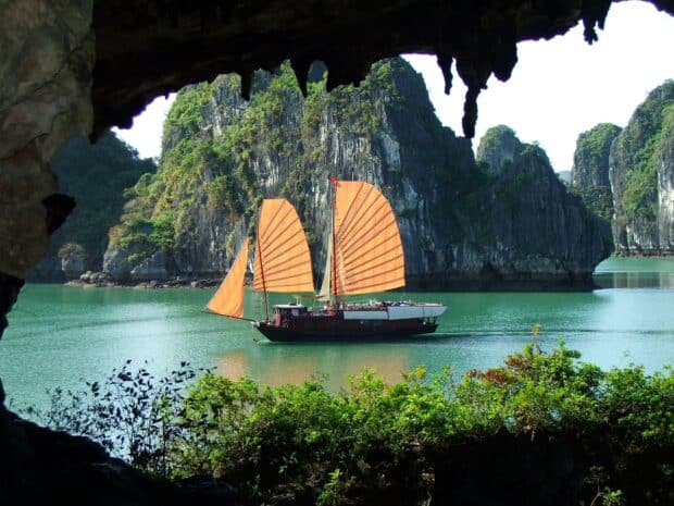 A traditional sailboat with orange sails floats on calm green waters surrounded by limestone cliffs in Vietnam, HD Desktop Wallpaper