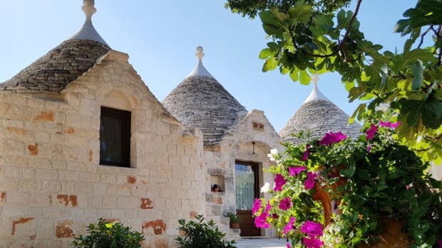 The photo features traditional trulli houses in Alberobello decorated with vibrant flowers and clear blue sky, HD Desktop Wallpaper