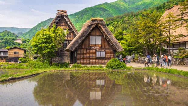 Traditional village houses reflect in a water filled rice field with people walking nearby in the village, HD Desktop Wallpaper