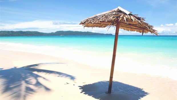 A beach umbrella stands on white sand near turquoise water with distant hills in the background, 4K Desktop Wallpaper