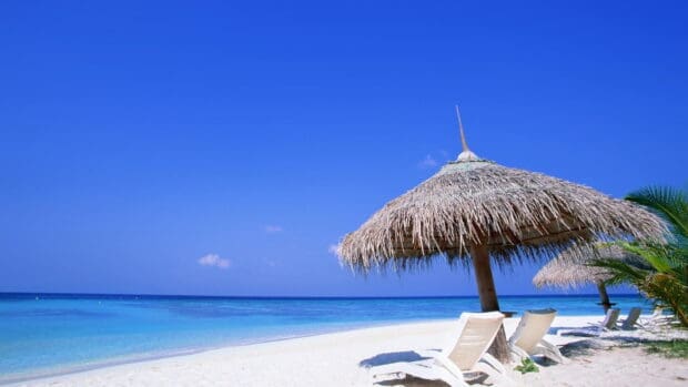 A tropical beach umbrella stands beside lounge chairs on the white sand near clear blue water, HD Desktop Wallpaper