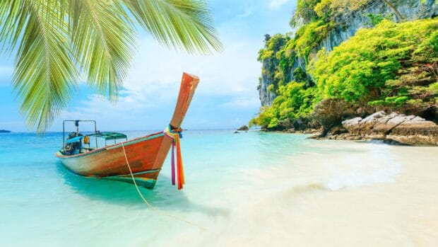 A wooden boat with colorful ribbons floats near a green island under a clear sky in Phuket, 2K Desktop Wallpaper