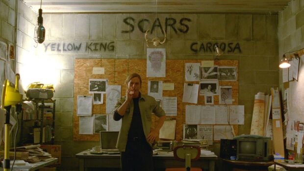 A detective stands thoughtfully in a cluttered room filled with notes and sketches about the Yellow King mystery, HD Desktop Wallpaper