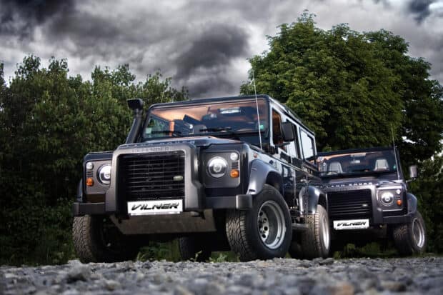 The image shows two Land Rover Defender vehicles parked outdoors near trees under a cloudy sky, 2K Desktop Wallpaper