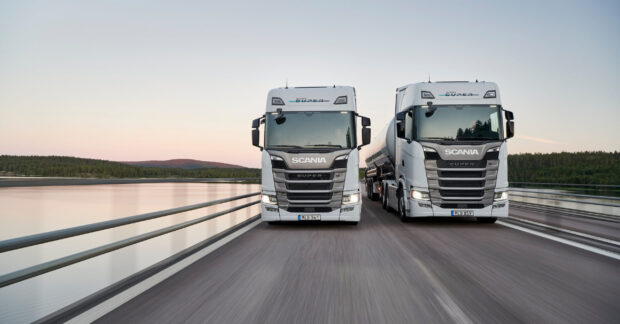 Two white Scania trucks are driving side by side on a bridge over water with a forest in the background featuring Scania, 2K Desktop Wallpaper