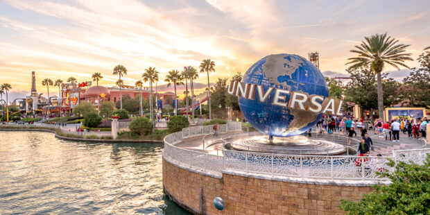 The Universal globe stands beside the water at sunset with palm trees and visitors around it in Orlando, 2K Desktop Wallpaper