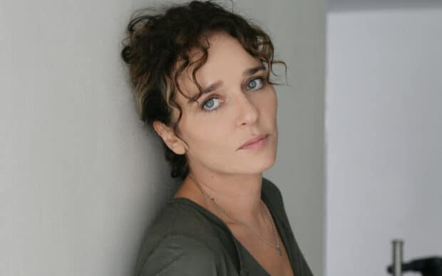 A close up of Valeria Golino looking thoughtfully at the camera leaning against a white wall, 2K Desktop Wallpaper