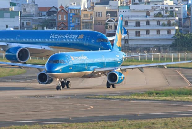 Two Vietnam Airlines jets are taxiing on the airport runway with city buildings in the background, HD Desktop Wallpaper