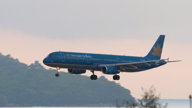 A Vietnam Airlines plane is flying low over water near a tree covered island at sunset, 4K Desktop Wallpaper