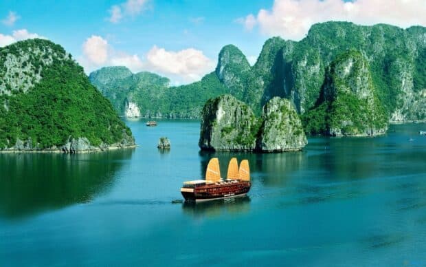 A traditional wooden boat sails through emerald waters surrounded by towering green limestone islands in Vietnam Wallpapers, 2K Desktop Wallpaper