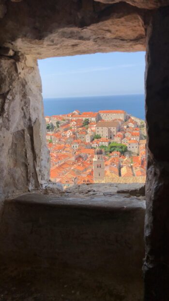 The ancient city walls of Dubrovnik are seen through a stone window with orange rooftops below, 4K Mobile Wallpaper